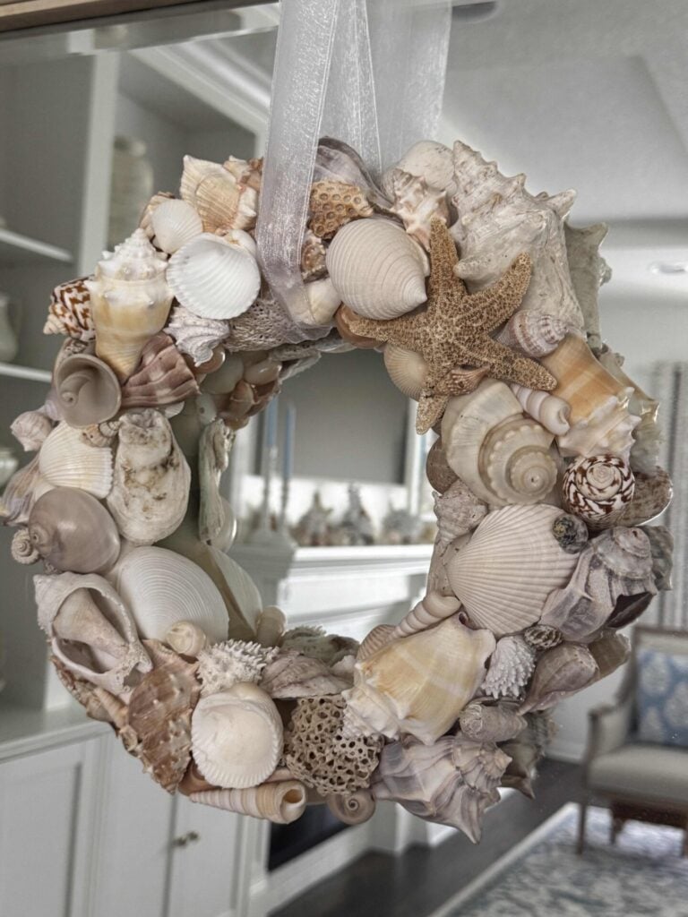 A DIY Shell Wreath made of assorted seashells and a starfish hangs by a sheer white ribbon indoors, with shelves and a mirror visible in the background.