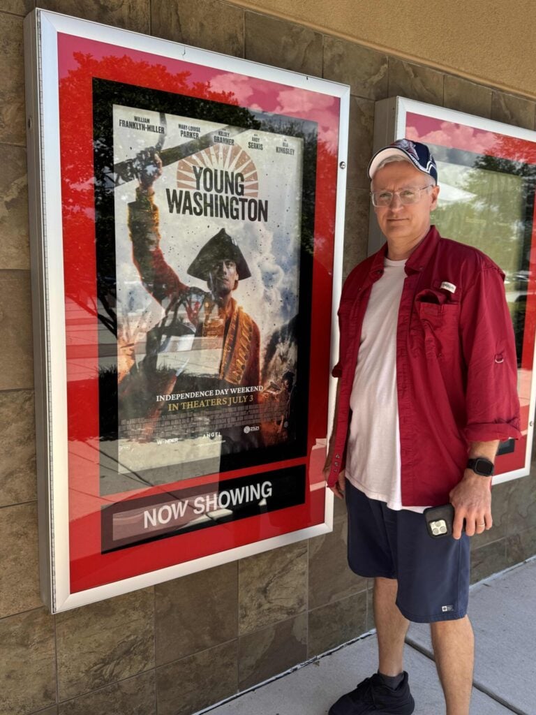 A man in a red shirt and cap stands next to a movie poster for Young Washington outside a theater-part for our Seasonal Sentiments July Rewind. A poster features a historical figure holding a flag. A Now Showing sign is visible below the poster.