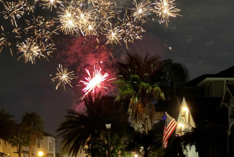 Fireworks light up the night sky above a suburban neighborhood with palm trees, parked cars, and a house displaying an illuminated American flag—a vibrant scene that captures the spirit of Seasonal Sentiments July 2026 Rewind.