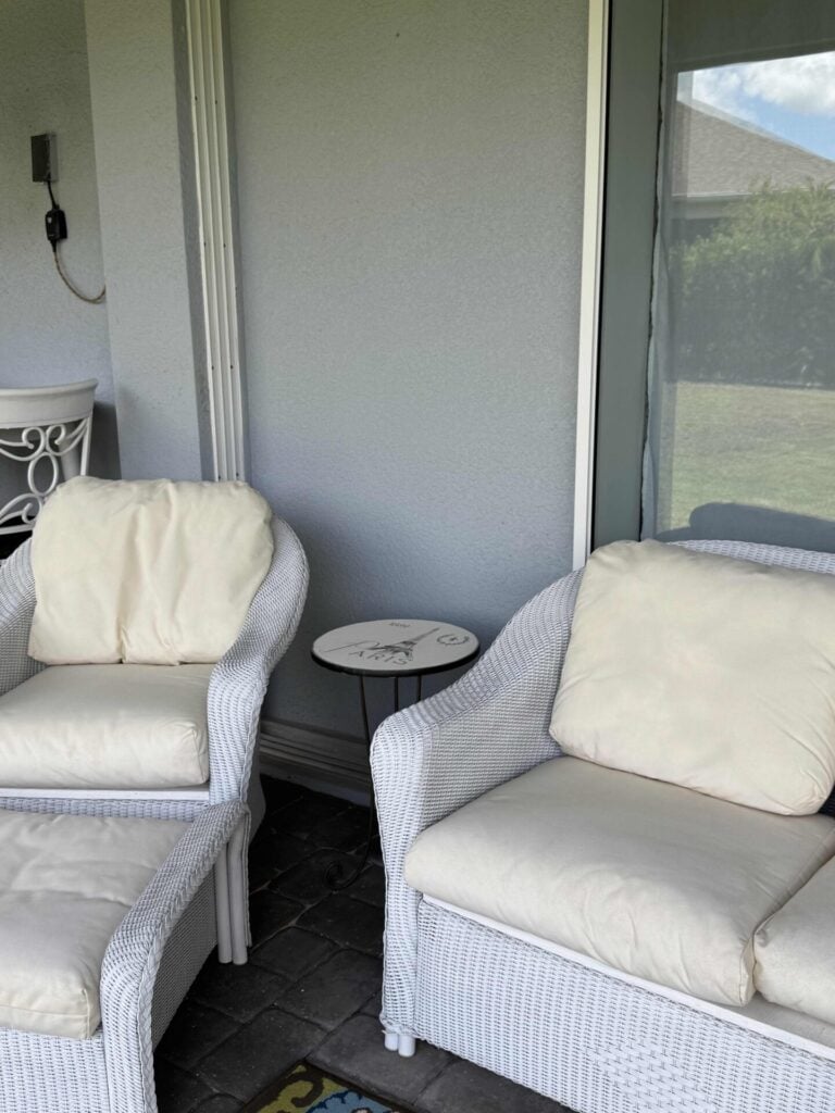 Two white wicker armchairs with cream cushions and matching ottomans sit on a patio next to a small round side table, placed in front of a glass door and window.