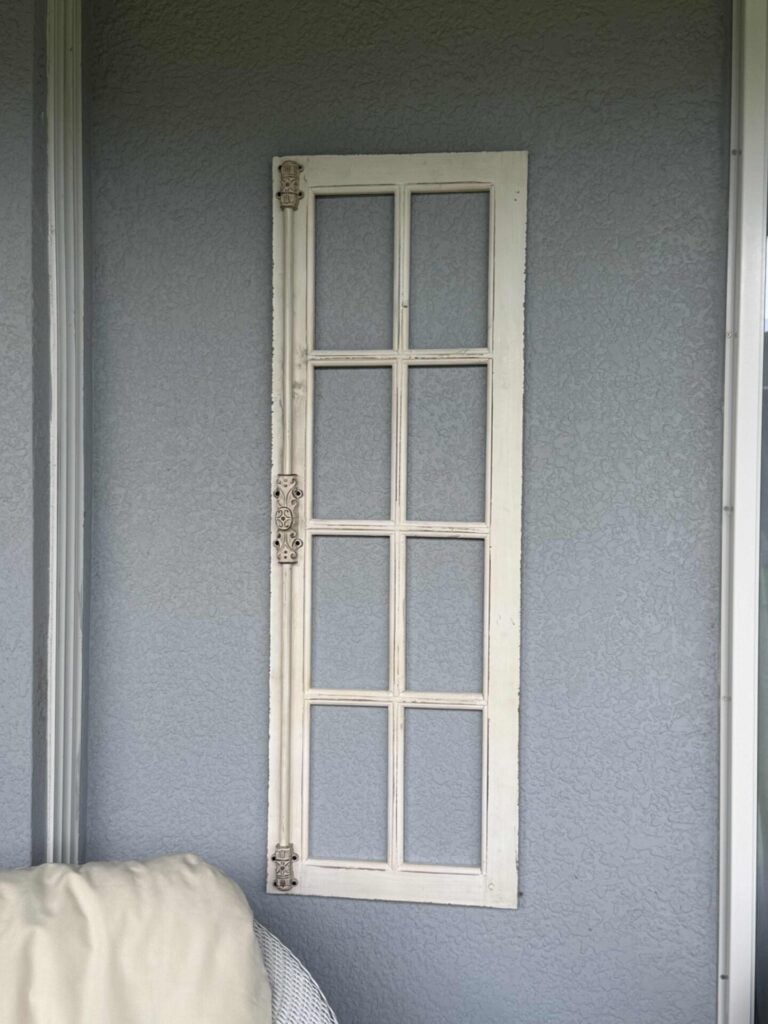 Step one for how to hang flower pots. A narrow, white, vintage-style window frame with six glass panes is mounted vertically on a textured gray wall. A beige cushion is partially visible in the lower left corner.