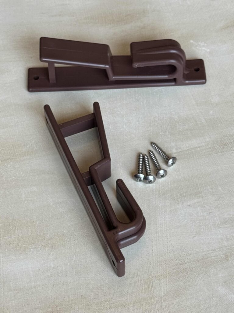 Two brown plastic wall brackets with three silver screws on a light textured surface. The brackets appear to be designed for holding rods or poles.