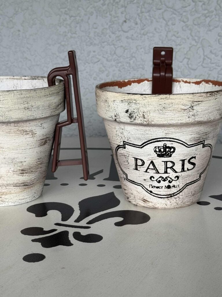 Two rustic white flower pots sit on a patterned surface. One pot says Paris Flower Market with a crown graphic, both have brown hooks attached. The background is light with a subtle textured pattern.