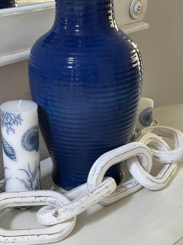 A large blue ceramic vase sits on a white dresser next to two white candles with blue nautical designs and a white decorative wooden chain.