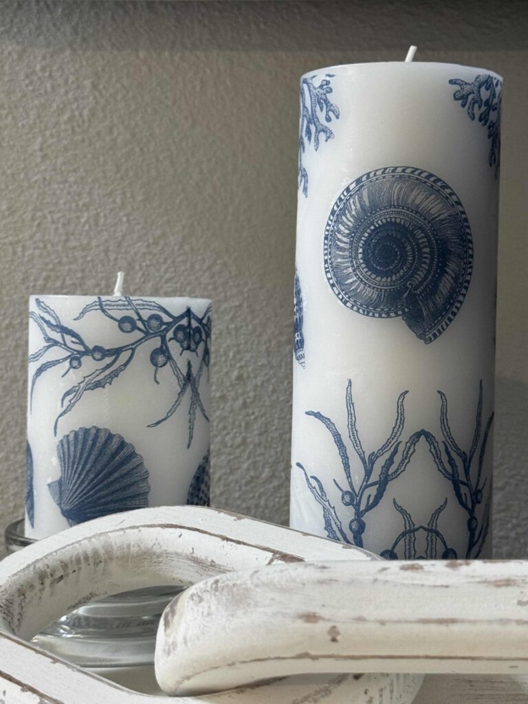 Two white pillar candles decorated with blue sea shells, seaweed, and marine designs sit on a reflective surface near a textured white wood accent piece against a neutral wall.
