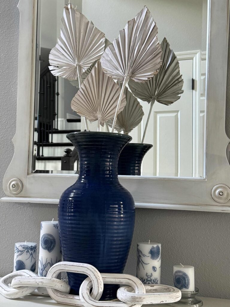 A blue ceramic vase with silver palm leaves sits in front of a mirror, surrounded by white decorative chain links and blue-and-white patterned candles on a gray surface—an elegant arrangement perfect for inspiring new Summer Decor Ideas.