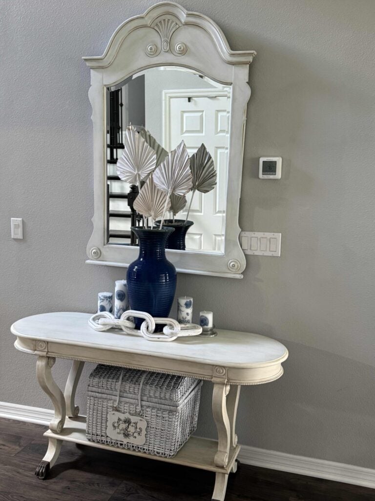 White console table with a blue vase holding three dried palm leaves—perfect for freshening up your space with subtle summer decor ideas. A large ornate mirror hangs above, reflecting natural light. Two small blue-and-white candles and a white woven basket sit on the table, all set against a soothing gray wall.