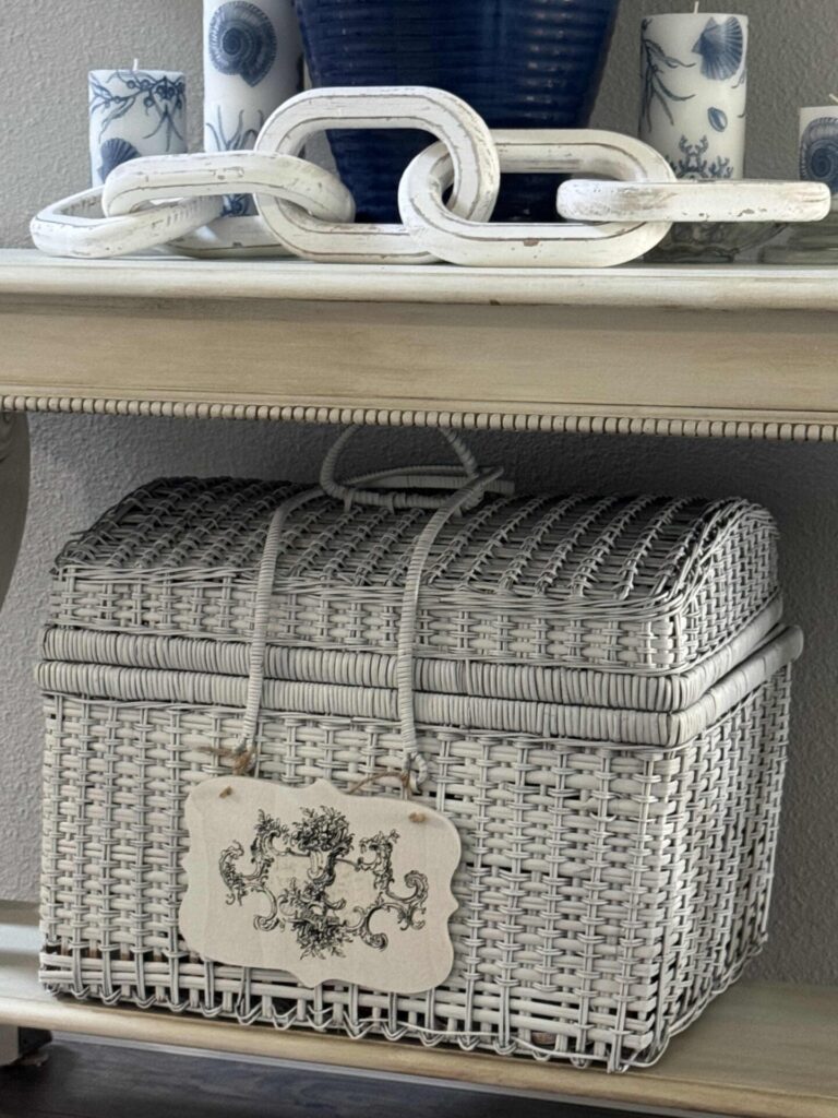 A white wicker basket with a rope handle and crest design sits on a shelf; above it are white chain links and blue and white vases, creating an inviting arrangement perfect for summer decor ideas.