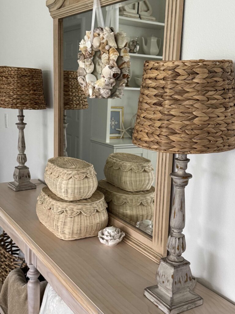 A wooden console table with wicker lamps, woven baskets, a seashell wreath hanging on a mirror, and a shell decor piece creates a coastal, rustic look—perfect for those seeking fresh Summer Decor Ideas.