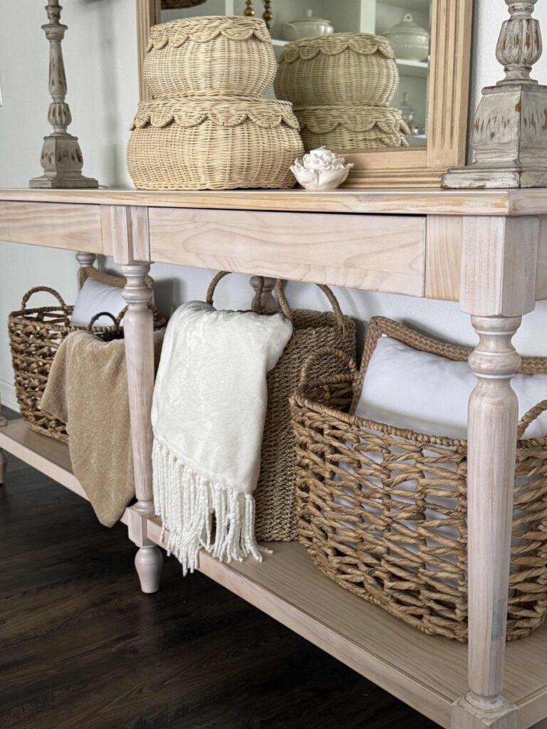 Light wood console table with decorative baskets, folded blankets, and pillows on the lower shelf. Woven baskets, a mirror, and sculpted decor sit on top.