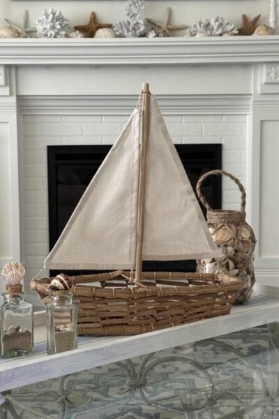 A woven sailboat with a canvas sail sits on a white table, surrounded by glass jars filled with seashells and a seashell-covered lantern—perfect Summer Decor Ideas for bringing coastal charm into your home. In the background, a mantel is decorated with white coral and starfish.