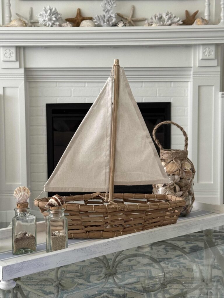 A woven sailboat with a canvas sail sits on a white table, surrounded by glass jars filled with seashells and a seashell-covered lantern—perfect Summer Decor Ideas for bringing coastal charm into your home. In the background, a mantel is decorated with white coral and starfish.