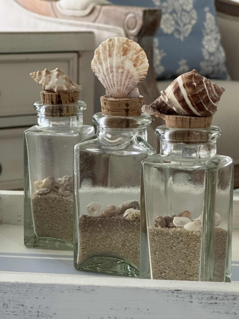 Three glass bottles filled with sand and small seashells are topped with corks, each decorated with a larger seashell. The bottles sit in front of a blurred background featuring furniture and a blue floral pillow.