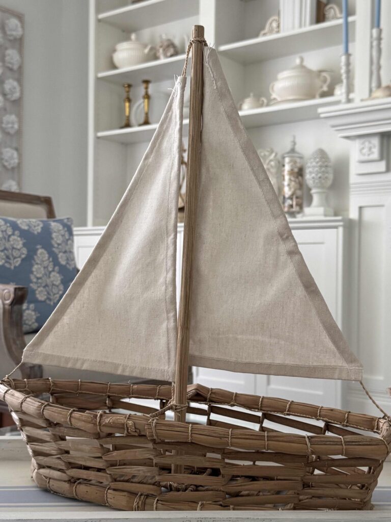 A woven wicker sailboat with beige fabric sails is displayed indoors on a table, while white shelves holding decorative ceramics and books create a relaxed, coastal backdrop.