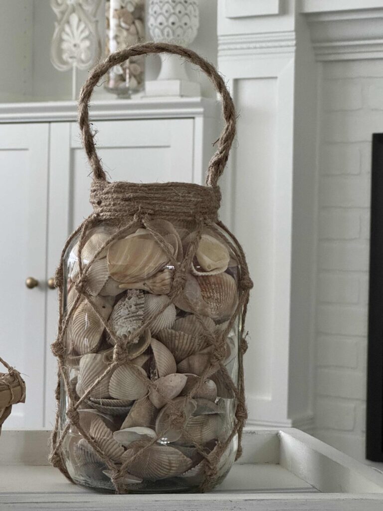 A glass jar filled with assorted seashells, wrapped in a rope net with a handle, sits on a white tray in a coastal-themed room.