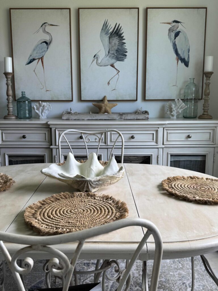 Coastal-themed dining room with a white table, four woven placemats, and a shell centerpiece. Three bird paintings hang above a white sideboard decorated with seashells, candles, and glass bottles—perfect for those seeking Summer Decor Ideas that bring the beachside charm indoors.