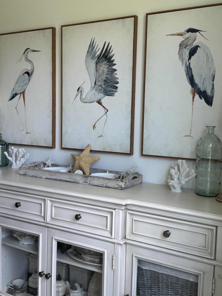 A white sideboard with coastal decor, including coral, glass bottles, and a starfish tray—perfect for summer decor ideas—sits beneath three framed bird paintings on a light-colored wall.