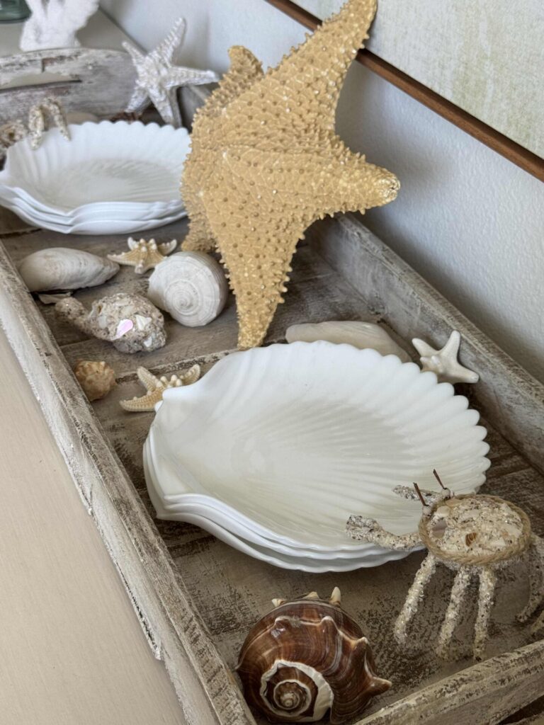 A wooden tray holds white scallop-shaped dishes, decorative starfish, seashells, and a crab figurine, creating a coastal-themed display on a light surface—perfect inspiration for summer decor ideas.
