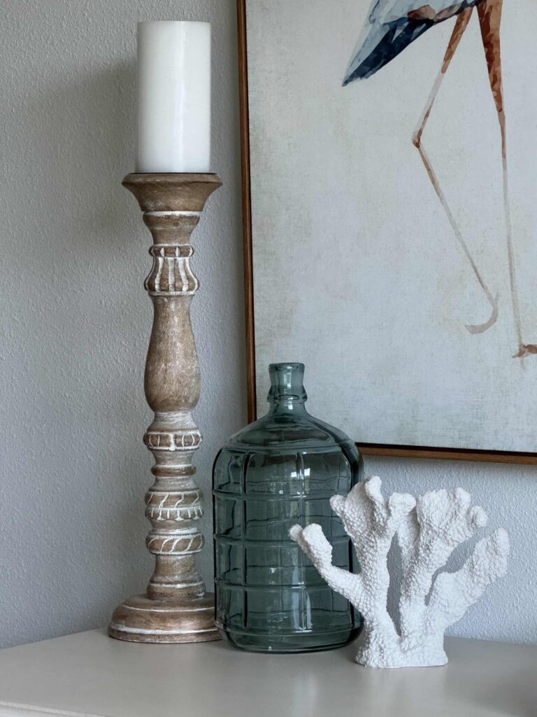 A wooden candle holder with a white candle, a clear glass jug, and a white coral decoration are arranged on a white surface next to a framed artwork featuring bird legs—perfect for inspiring Summer Decor Ideas with their fresh, coastal charm.