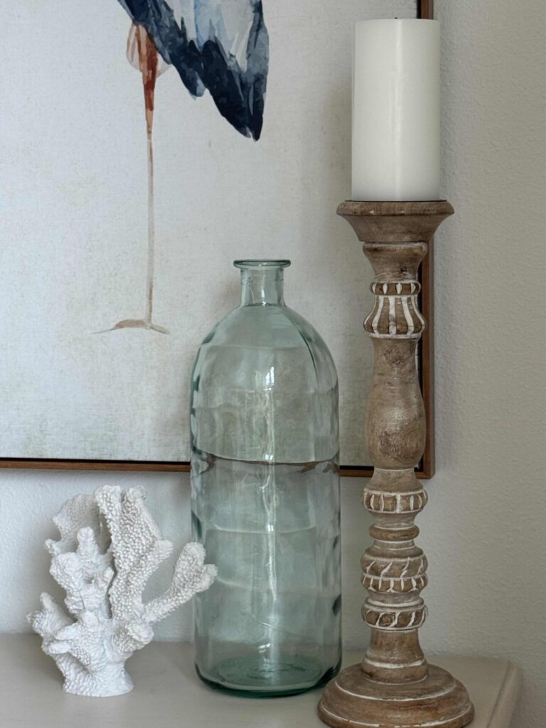 A decorative arrangement with a white candle on a wooden holder, a clear glass bottle, and a white coral ornament—perfect for summer decor ideas—is set before a framed painting with a bird’s leg visible.