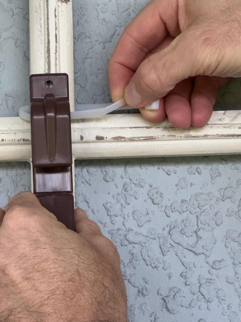 Step two for how to hang flower pots. Two hands secure a brown plastic bracket to a white wooden frame using a zip tie, against a textured light blue wall.
