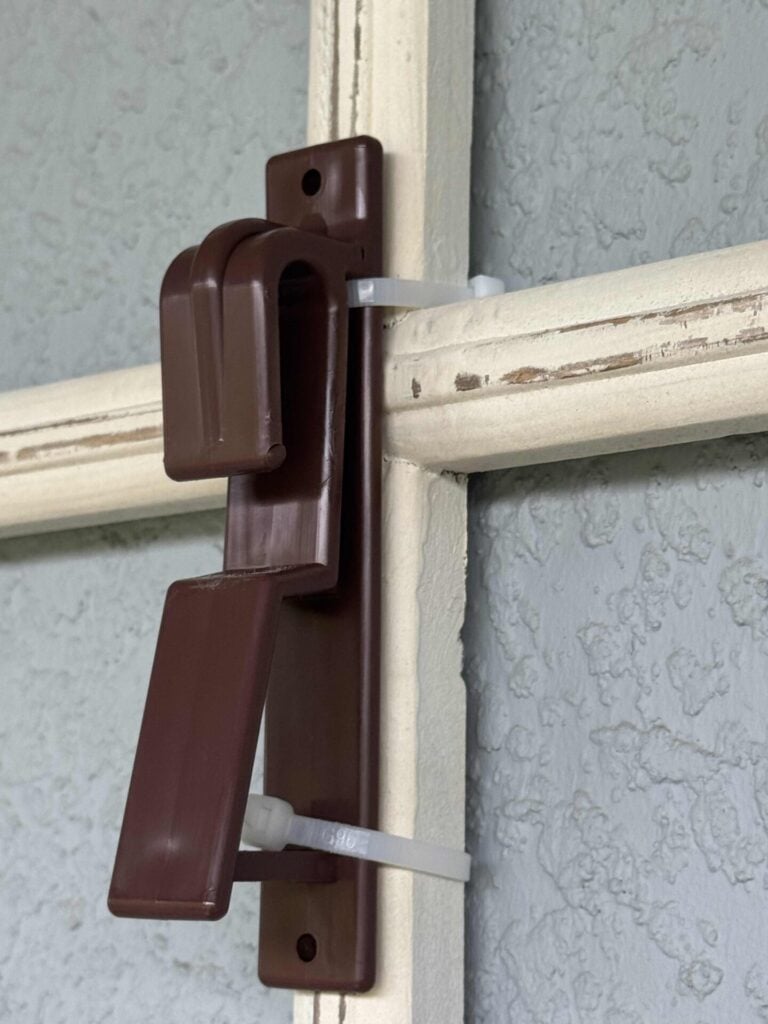 A brown plastic door latch is attached to a window frame using white zip ties instead of screws. The frame is cream-colored and the wall behind is textured and light blue.