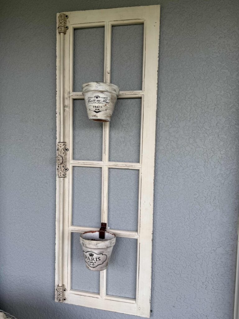 Step three for how to hang flower pots. A vintage white window frame hangs vertically on a gray textured wall, holding two distressed white flower pots labeled “PARIS,” one in the top section and one in the bottom section.
