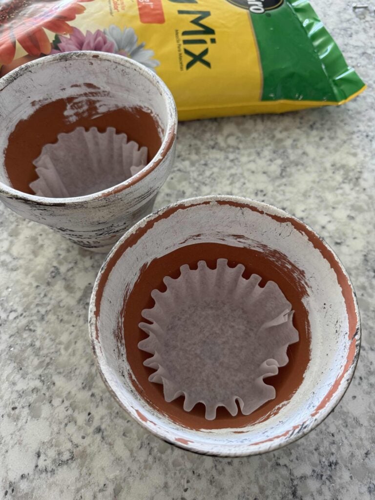Step four for how to hang flower pots. Two empty terracotta pots with white paint on the edges, each lined with a coffee filter, sit on a speckled countertop. A bag of potting mix is partially visible in the background.