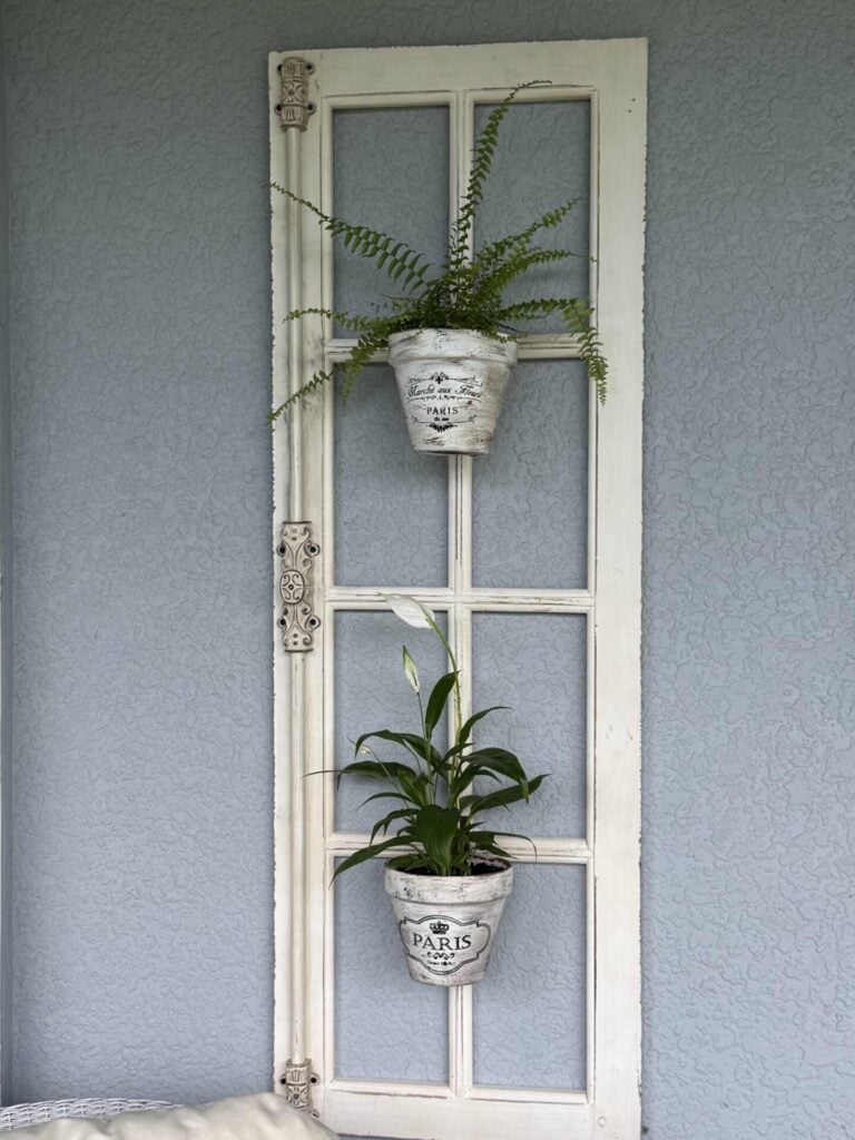 A vintage white window frame is mounted on a blue wall with two potted plants hanging on it. The pots are white with black Paris-themed text and designs. The upper plant is a fern, and the lower plant has long, broad leaves.