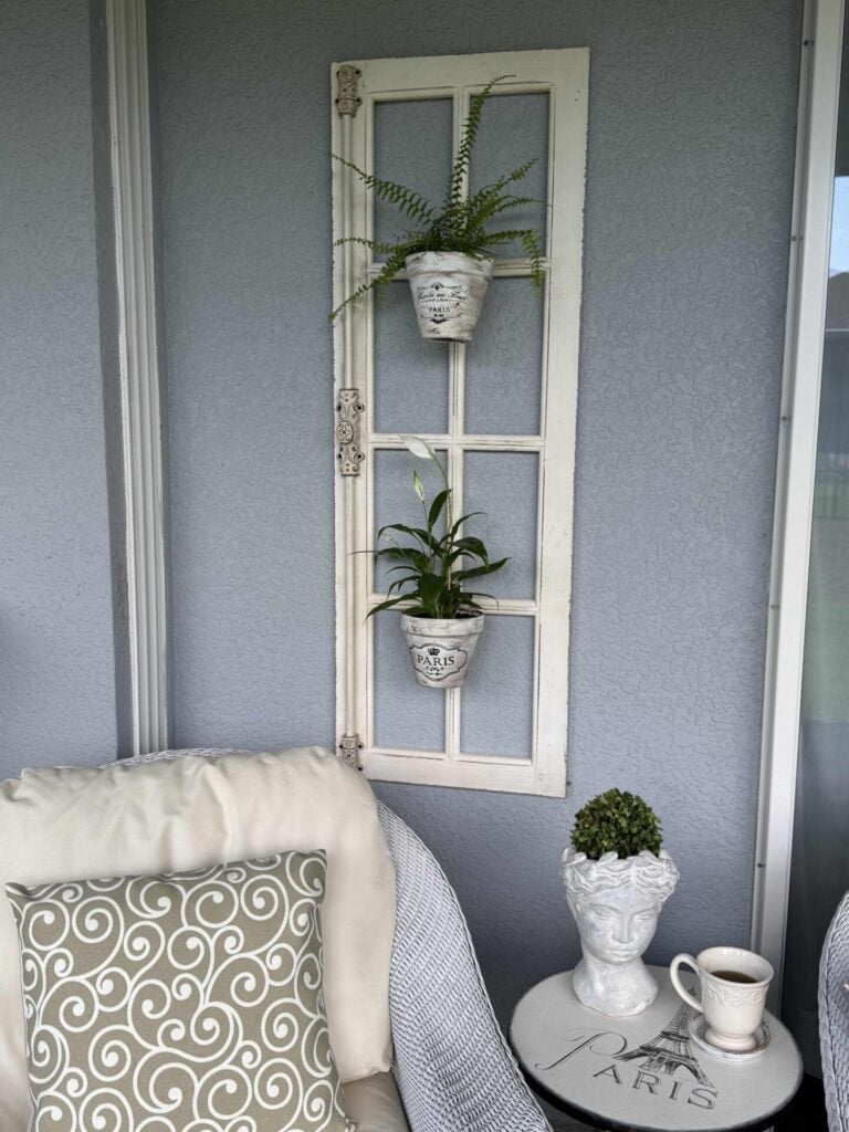 A cozy patio corner with a white chair, swirl-patterned pillow, side table with a cup, decor, and a vintage window frame on the wall holding two potted plants.