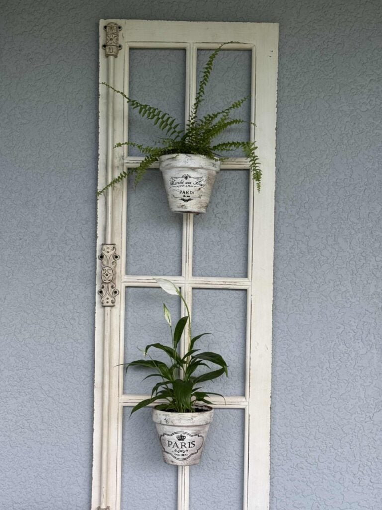Step five for how to hang flower pots. A vintage white window frame hangs on a gray textured wall, decorated with two potted plants in white pots labeled Paris, one fern at the top and another leafy green plant below.