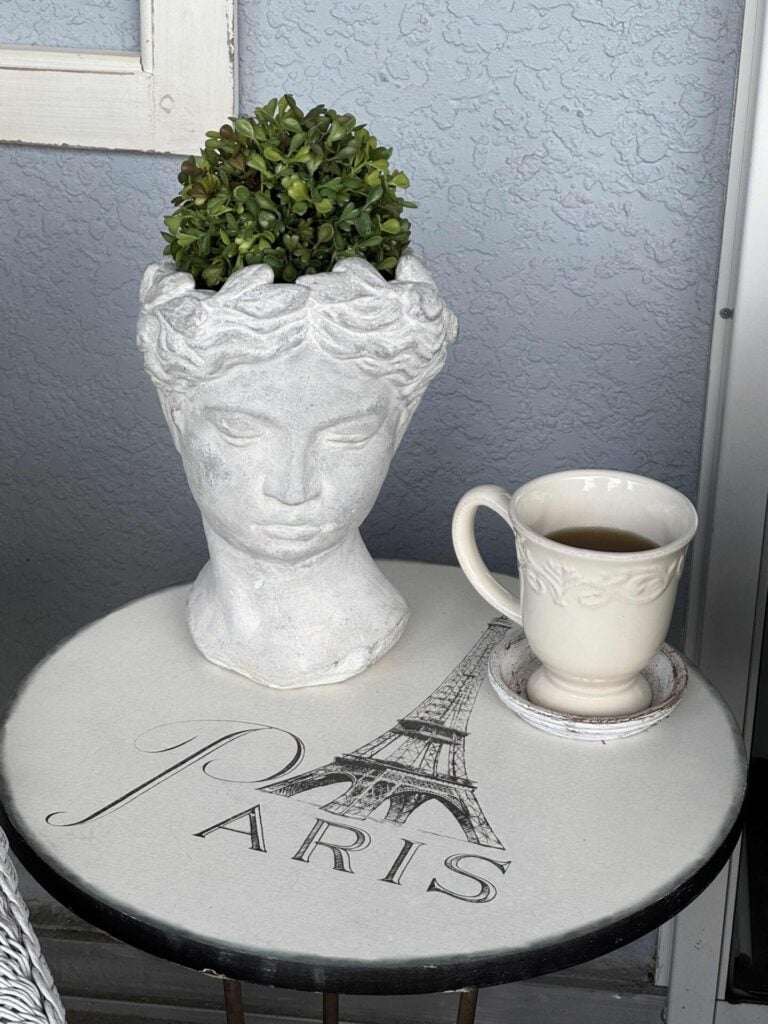 A white bust planter with green foliage and a cup of tea sit on a round table decorated with the Eiffel Tower and the word “Paris” against a light blue textured wall.