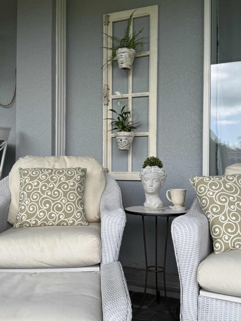 Two white wicker chairs with swirl-patterned pillows sit by a small table holding a bust planter and a mug. An old window frame with potted plants hangs on the gray wall behind them.