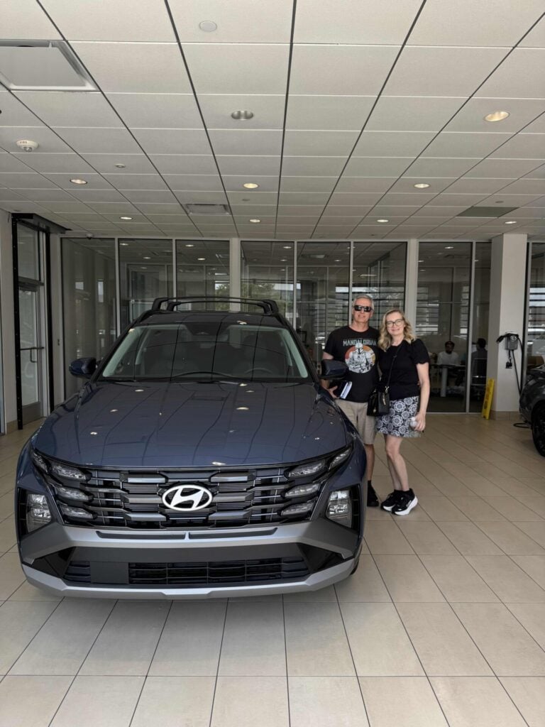 Two people stand smiling next to a new blue Hyundai SUV inside a car dealership showroom with glass walls and tiled floors.