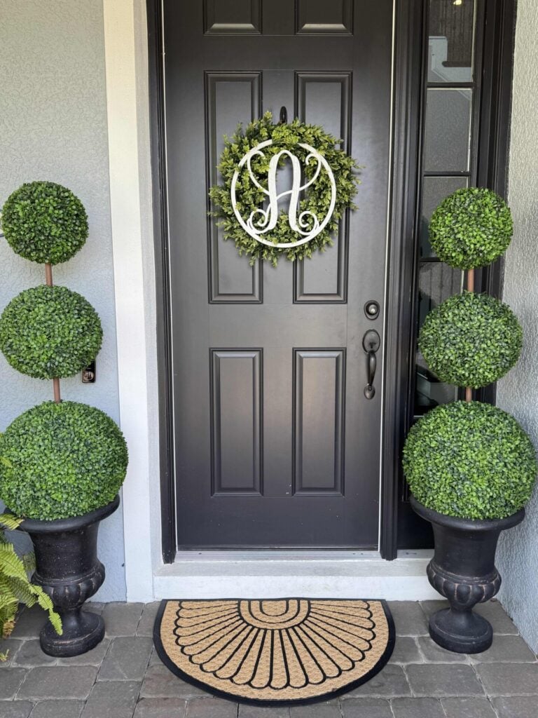 A black front door with a green wreath featuring a white script A creates a welcoming entry. Two tall DIY topiary trees in black pots flank the door, adding an elegant, personalized touch. A tan semicircle doormat sits on gray pavers in front of the entrance.
