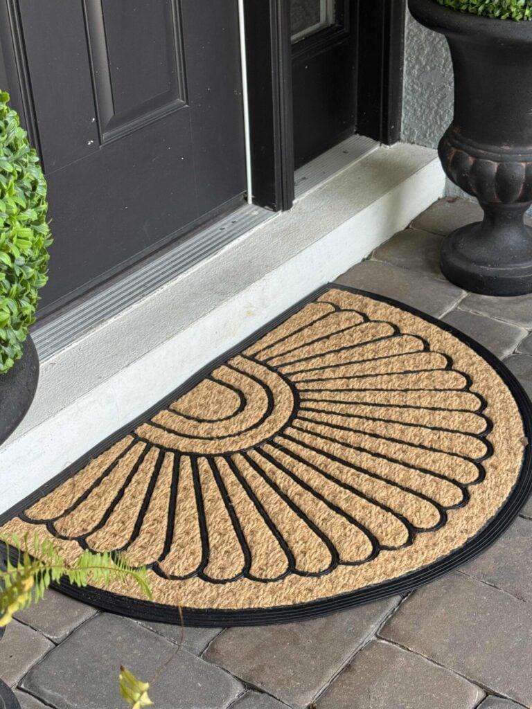 A semicircle doormat with a black sunburst pattern lies in front of a black door, surrounded by potted plants and gray stone tiles.