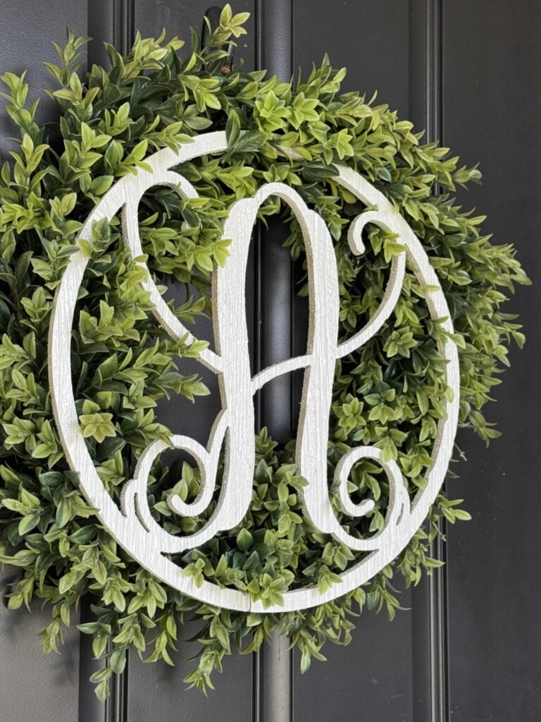 A green leaf wreath hangs on a dark door, featuring a large white letter A in the center, made from a decorative script font.