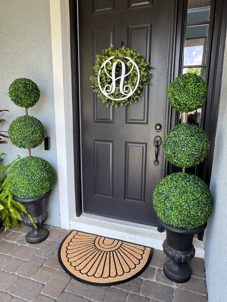 A dark front door with a green wreath displaying a white monogram A welcomes guests. On each side, DIY topiary trees in black pots add a touch of elegance, while a tan semicircle doormat lies on the stone-tiled porch.