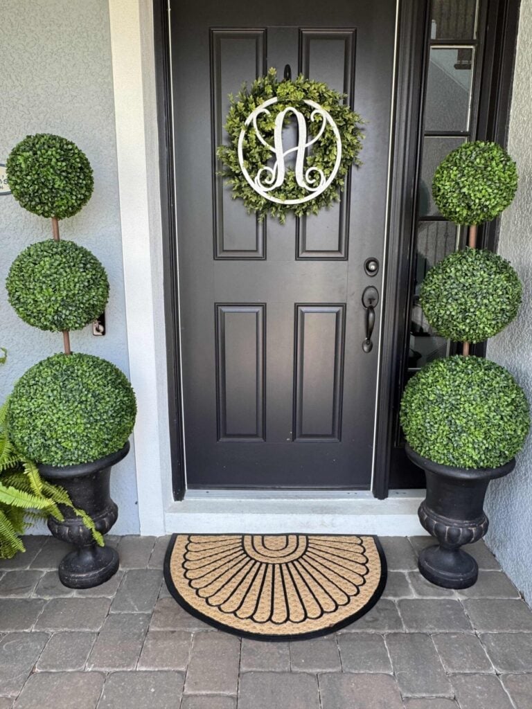A black front door with a green leafy wreath featuring a white letter A is flanked by two DIY topiary trees in pots and a semicircle doormat on stone paving.