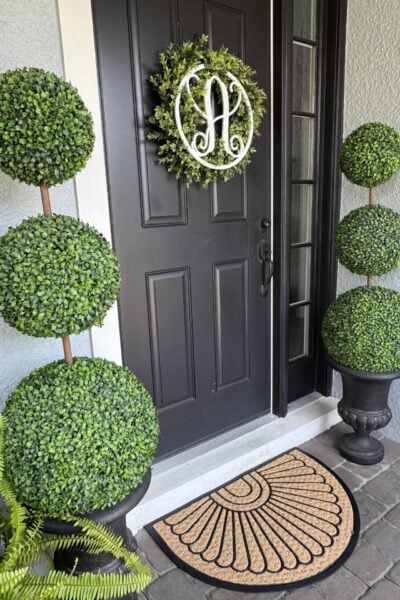 Black front door with a circular green wreath featuring a white letter A. On either side, two DIY topiary trees in black pots add architectural charm, while a tan doormat with a semicircle pattern sits at the doorstep.