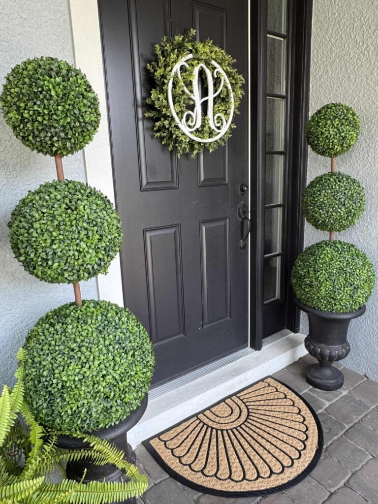 Black front door with a circular green wreath featuring a white letter A. On either side, two DIY topiary trees in black pots add architectural charm, while a tan doormat with a semicircle pattern sits at the doorstep.