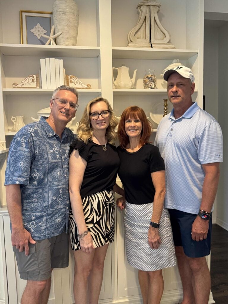 Four adults stand together smiling in front of white shelves with decorative items. Two women are in the center, each flanked by a man. They are casually dressed and appear to be posing for a group photo indoors.