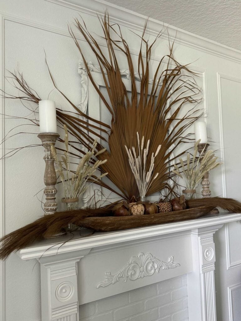 A white fireplace mantel is decorated with tall dried palm leaves, pampas grass, wooden candlesticks with white candles, pinecones, and other natural elements, creating a rustic, earthy vibe.