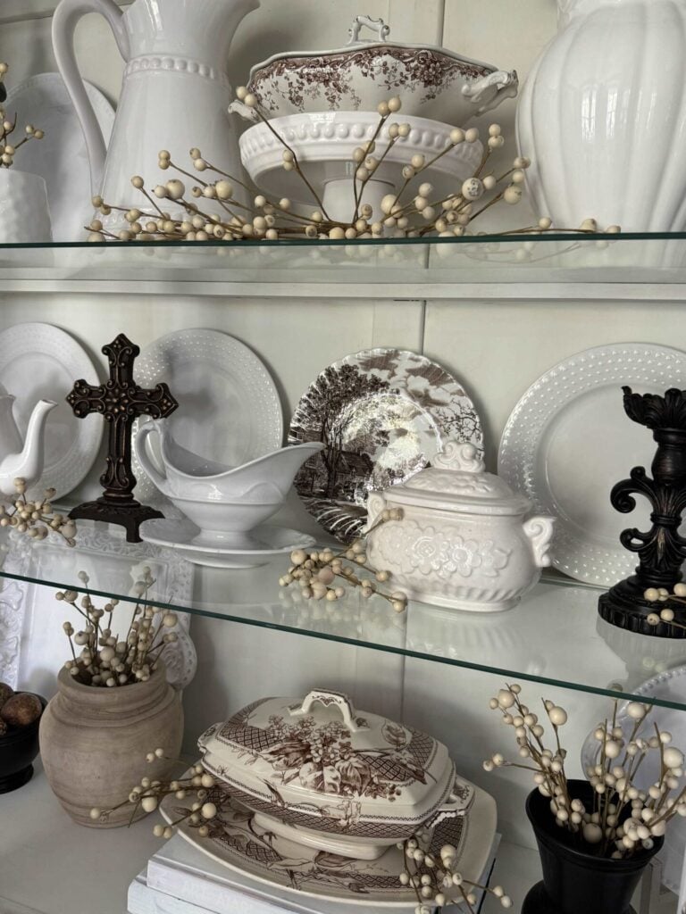 Glass shelves display white and brown vintage dishes, ornate pitchers, plates, teapots, and serving bowls, accented by sprigs of white berries and decorative black cross and urn pieces.