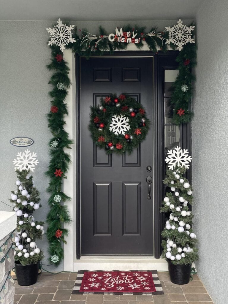 A black front door decorated with a green wreath, garland with red bows and white snowflakes, two DIY Topiary Trees adorned with white ornaments, and a red “Merry Christmas” doormat.