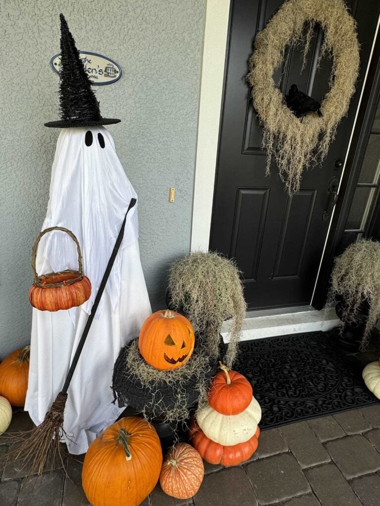 A ghost figure with a witch hat, broom, and pumpkin basket stands by a black door decorated with a wreath. Stacked pumpkins and moss surround the ghost, creating a festive Halloween scene.