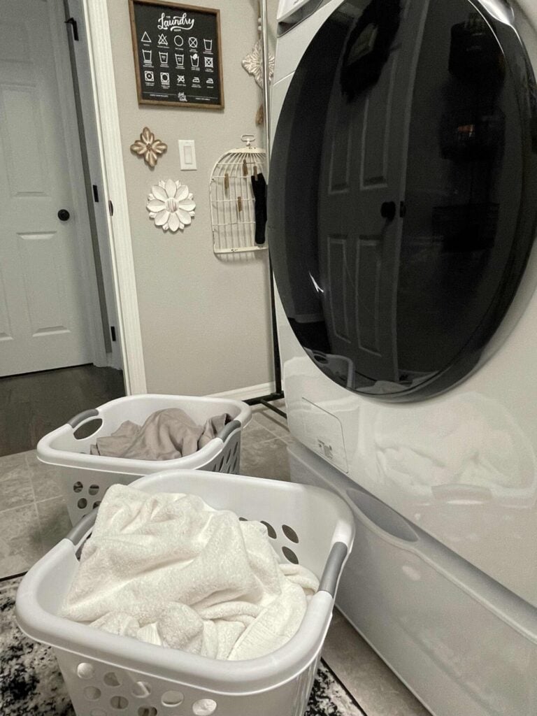 Two laundry baskets with clothes sit on the floor next to a front-loading washing machine in a laundry room decorated with wall art and a rug.