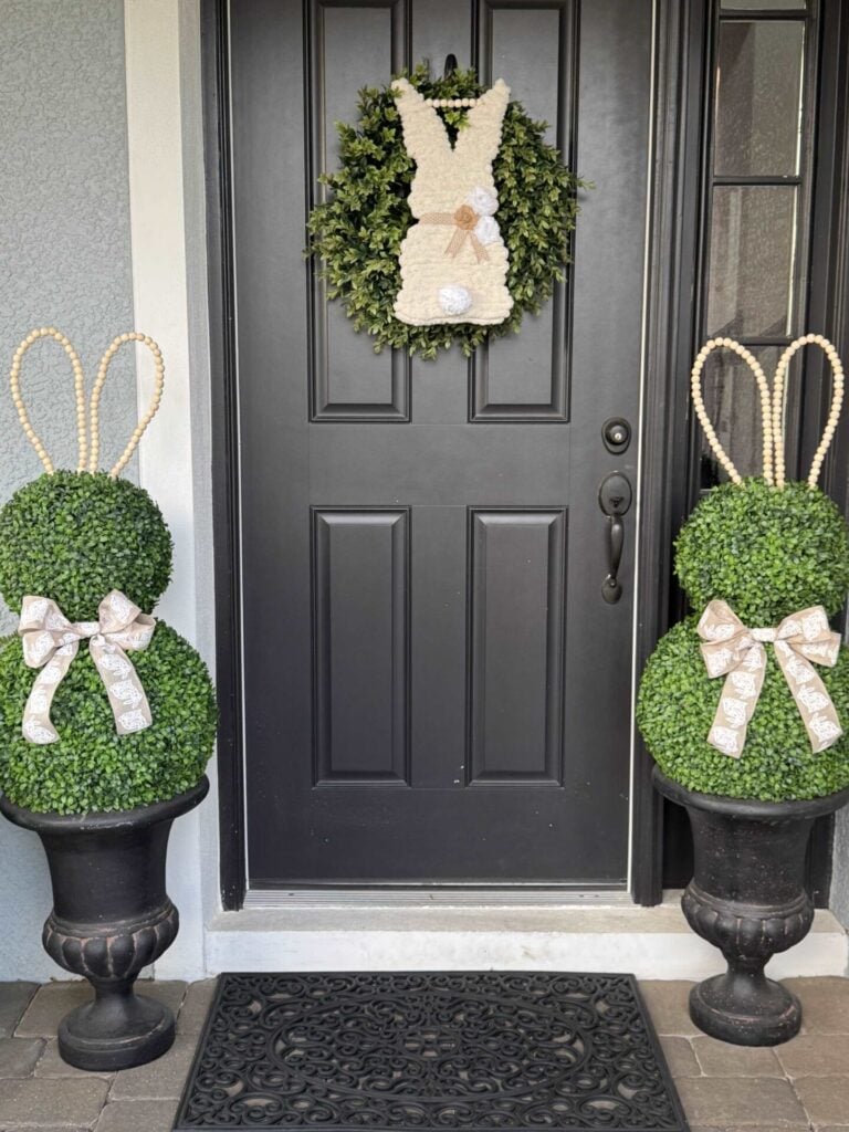 Two DIY Topiary Tree bunny figures with bows stand in urns on each side of a black door, which features a wreath with a white bunny decoration at its center. A black doormat sits in front of the door.