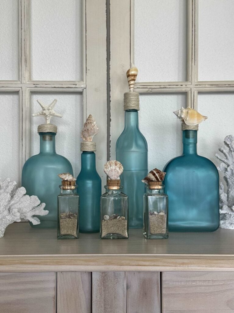 A display of blue and clear glass bottles of various sizes on a wooden surface, each topped with seashells, evokes coastal charm. Some bottles contain sand and small shells, while white coral decor and a DIY Shell Wreath complete the look by the window.