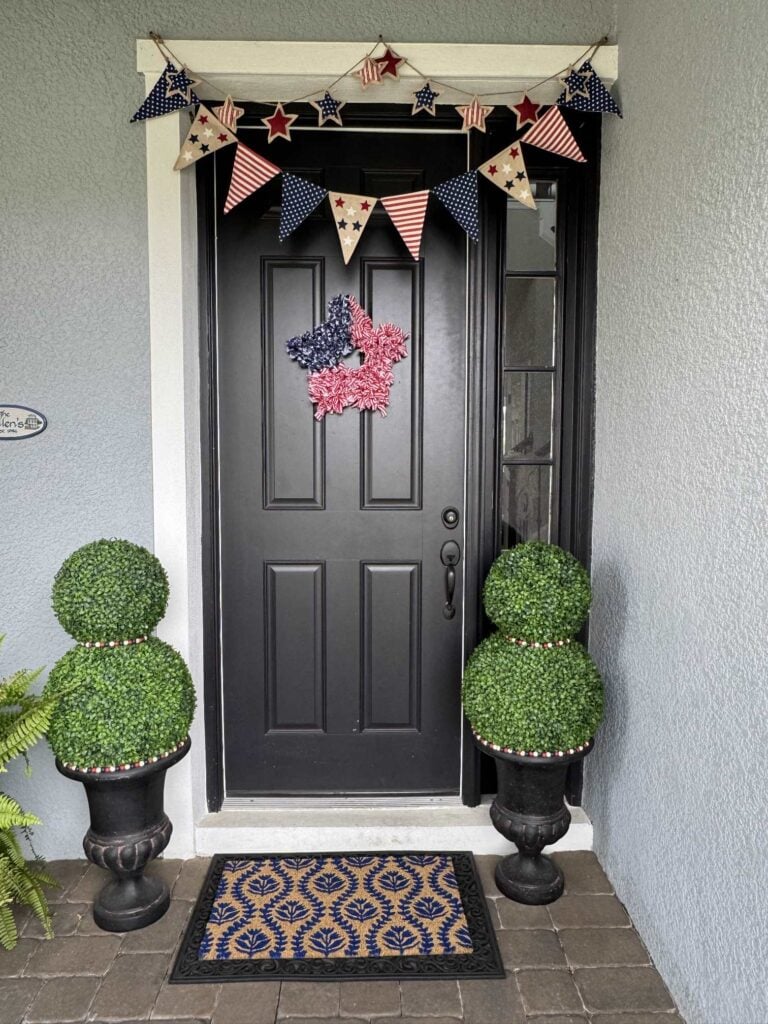 A black front door with patriotic red, white, and blue star and flag banners, a pink star wreath, two DIY topiary trees in pots, and a doormat with a blue leaf pattern.